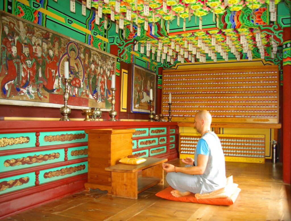 Michael G. Leitner sitzt meditierend auf einem Kissen in einem farbenprächtig gestalteten buddhistischen Tempel mit traditionellen Wandmalereien und hängenden Laternen.