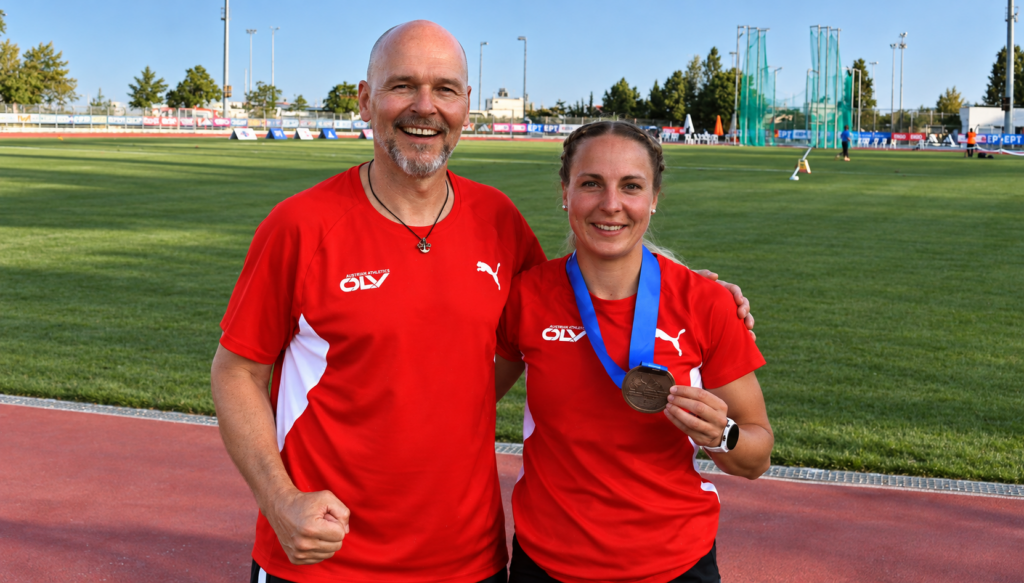 Michael G. Leitner mit einer österreichischen Leichtathletin im ÖLV-Nationalteam-Outfit auf einem Leichtathletikplatz. Beide lächeln, die Sportlerin präsentiert ihre Medaille.