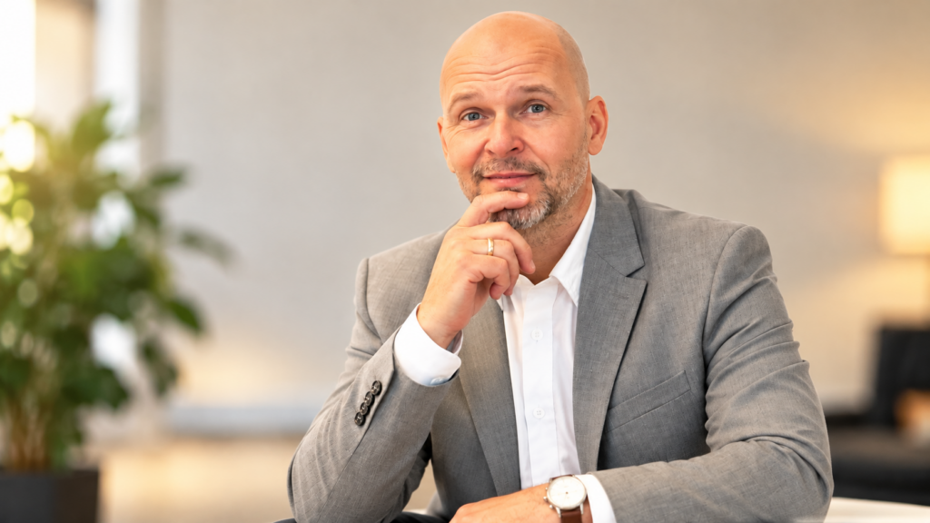 Michael G. Leitner, Mentalcoach für Business und Sport, sitzt in einem grauen Sakko mit nachdenklichem Blick und einer Hand am Kinn in einem hellen Büro. Kopieren
